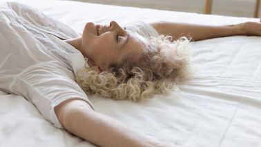 Frau liegt mit ausgestreckten Armen auf dem Bett – Symbol für entspannenden Schlafkomfort