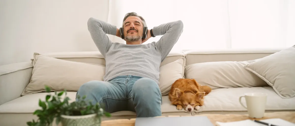 Älterer Mann sitzt mit Hund auf Couch und hört Musik zum Entspannen.