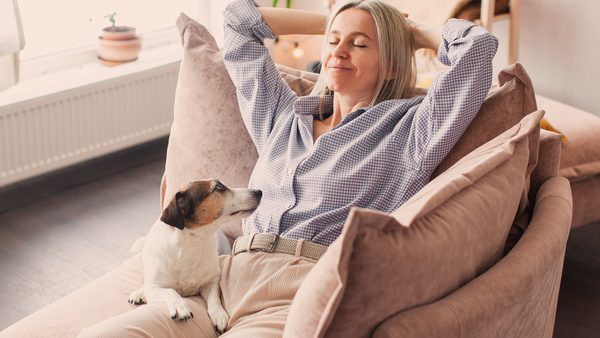 Frau mittleren Alters liegt entspannt auf dem Sessel auf ihrem Schoß sitzt ein Hund