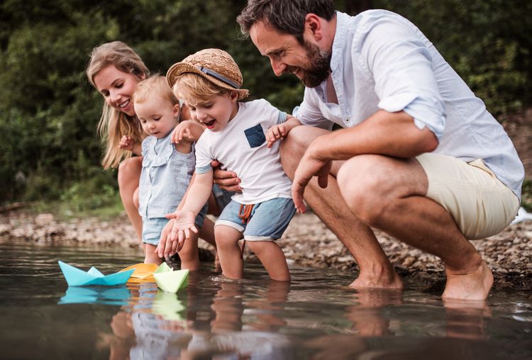 Familie mit zwei kleinen Kindern spielt am Wasser und lässt Papierboote auf dem Wasser fahren