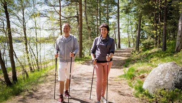 Zwei Personen beim Nordic Walking auf einem sonnigen Waldweg, umgeben von grünen Bäumen – ideal für gelenkschonendes Training und starke Knie