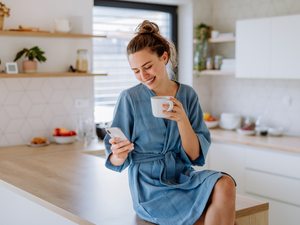 Frau in der Küche mit Tasse und Smartphone, entspannt im Umgang mit Unterleibsschmerzen während der Periode.