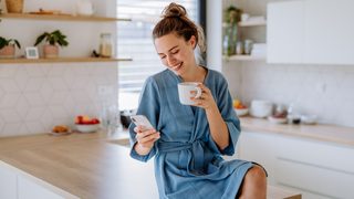 Frau in der Küche mit Tasse und Smartphone, entspannt im Umgang mit Unterleibsschmerzen während der Periode.