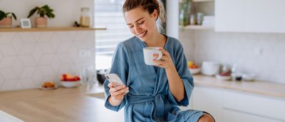 Frau in der Küche mit Tasse und Smartphone, entspannt im Umgang mit Unterleibsschmerzen während der Periode.