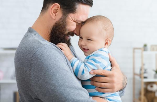 Ein Baby lacht in den Armen seines Vaters. Geborgenheit und Nähe fördern einen entspannten Schlaf für unruhige Babys.