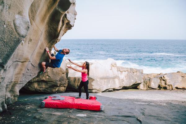 Mann bouldert an einem Felsen am Strand. Frau gibt Hilfestellung.
