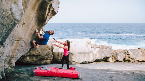 Mann bouldert an einem Felsen am Strand. Frau gibt Hilfestellung.
