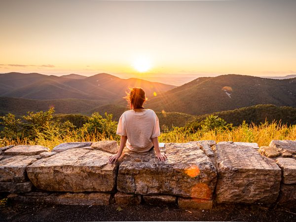 Person sitzt bei Sonnenuntergang auf einer Steinmauer und blickt über Berge – Symbol für Entlastung und Ruhe bei Hämorrhoiden-Beschwerden.