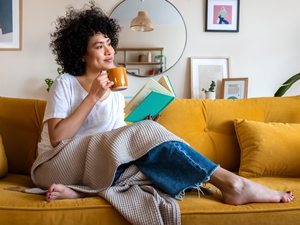 Frau auf gelbem Sofa mit Buch und Tee – Entspannung und Umgang mit PMS-Symptomen.