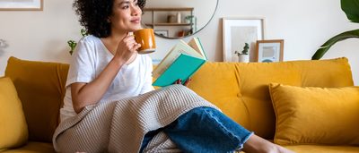 Frau auf gelbem Sofa mit Buch und Tee – Entspannung und Umgang mit PMS-Symptomen.