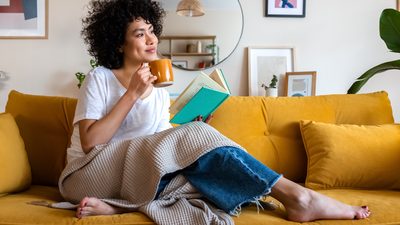 Frau auf gelbem Sofa mit Buch und Tee – Entspannung und Umgang mit PMS-Symptomen.