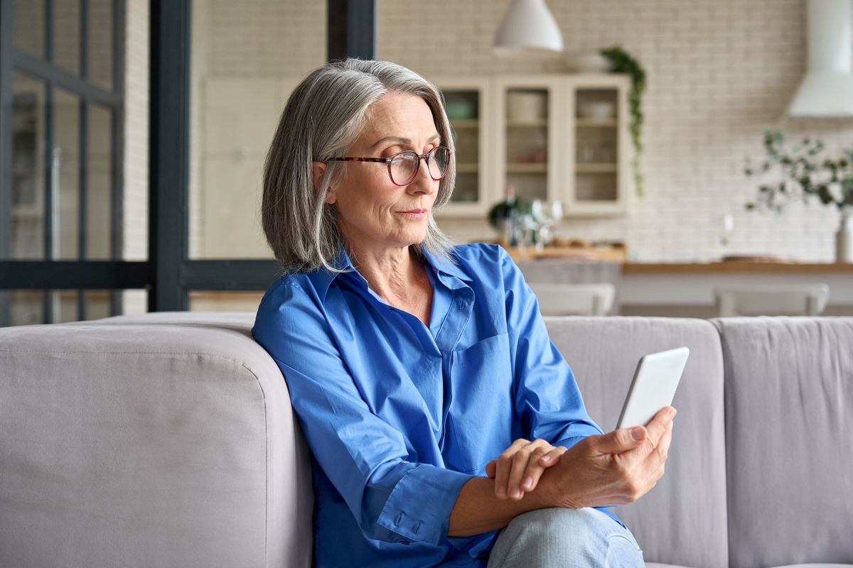 Ältere Frau mit Handy in der Hand auf einer Couch.