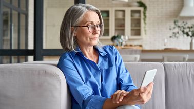 Ältere Frau mit Handy in der Hand auf einer Couch.