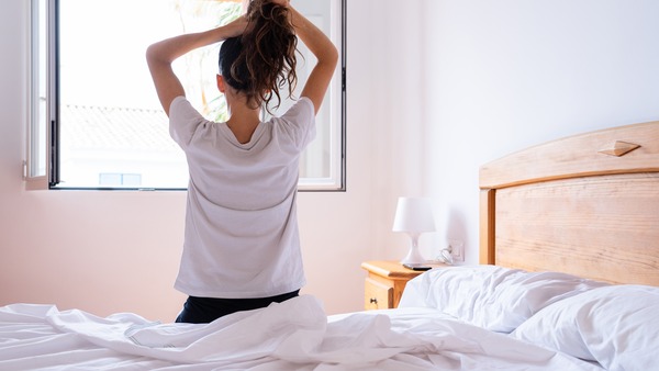 Frau sitzt auf ihrem Bett am offenen Fenster und bindet sich die Haare zusammen - Symbol für einen entspannten Start in den Tag