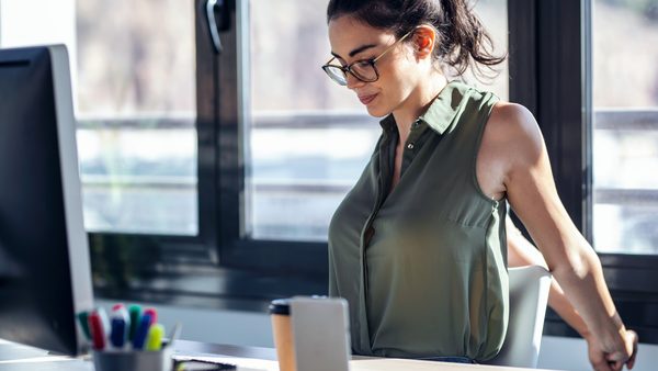 Frau sitzt am Schreibtisch und dehnt ihre Arme hinter dem Rücken.