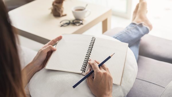 Eine Frau sitzt entspannt auf ihrem Sofa und schreibt in ihr Tagebuch