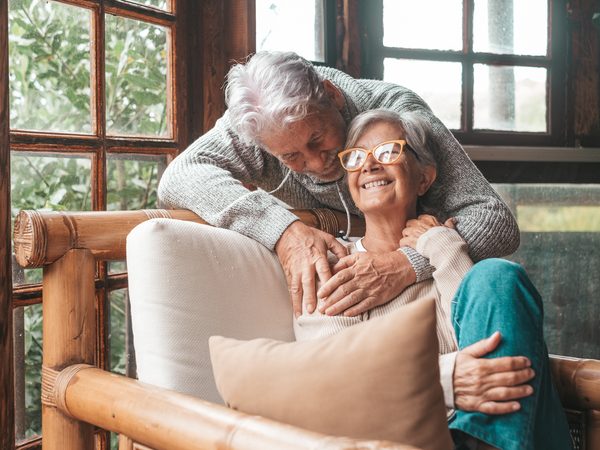 Ältere Frau sitzt auf einem Sessel und wird von ihrem Mann von hinten umarmt