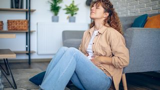 Frau sitzt mit einem schmerzverzerrten Gesicht und den Händen auf dem Bauch auf dem Boden