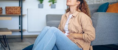 Frau sitzt mit einem schmerzverzerrten Gesicht und den Händen auf dem Bauch auf dem Boden