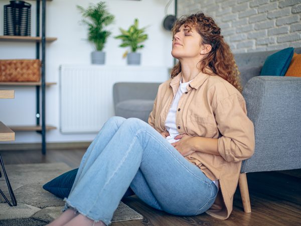 Frau sitzt mit einem schmerzverzerrten Gesicht und den Händen auf dem Bauch auf dem Boden