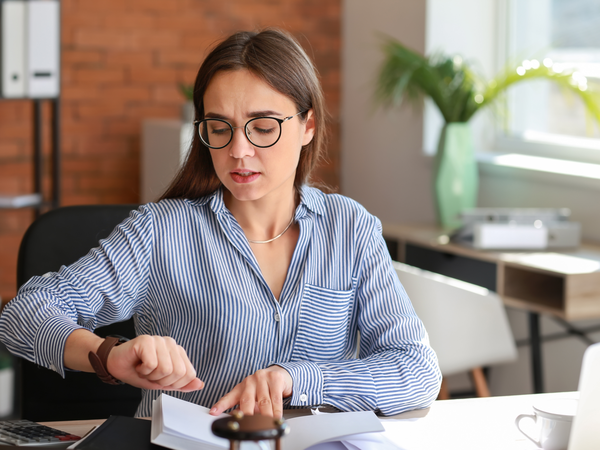 Gestresste Frau schaut am Schreibtisch auf die Uhr – Zeitdruck kann nervöse Herzbeschwerden begünstigen