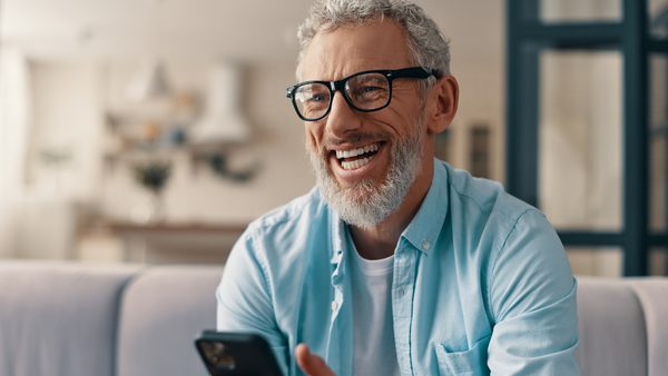 Stress durch soziale Medien: Mann mit Brille sitzt lächelnd auf dem Sofa und nutzt sein Smartphone zu Hause.