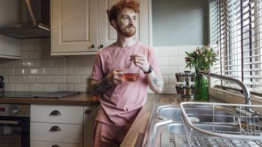 Mann lehnt sich in der Küche an, hat eine Tasse Tee in der Hand und schaut aus dem Fenster