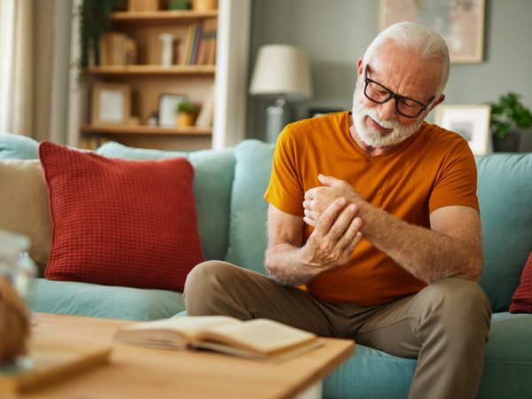 Älterer Mann in orangefarbenem T-Shirt sitzt auf einem Sofa und hält sein schmerzendes Handgelenk.
