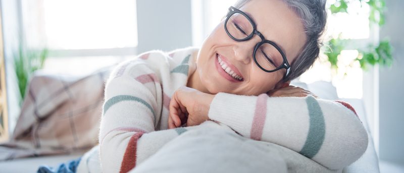 Frau mit Brille lächelt entspannt auf dem Sofa – kurze Ruhepause als sanfte Entlastung bei Kopf- & Nervenschmerzen.