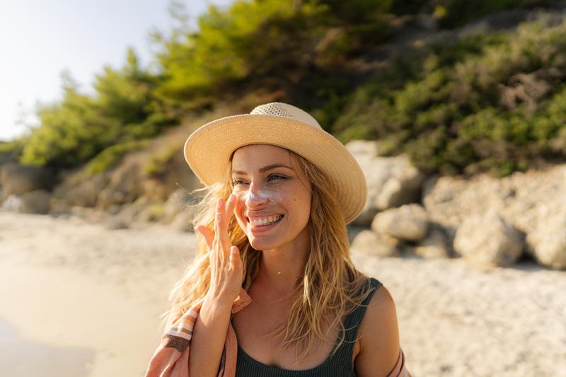 Frau mit einem Sonnenhut steht in der Sonne und cremt sich mit Sonnencreme ein