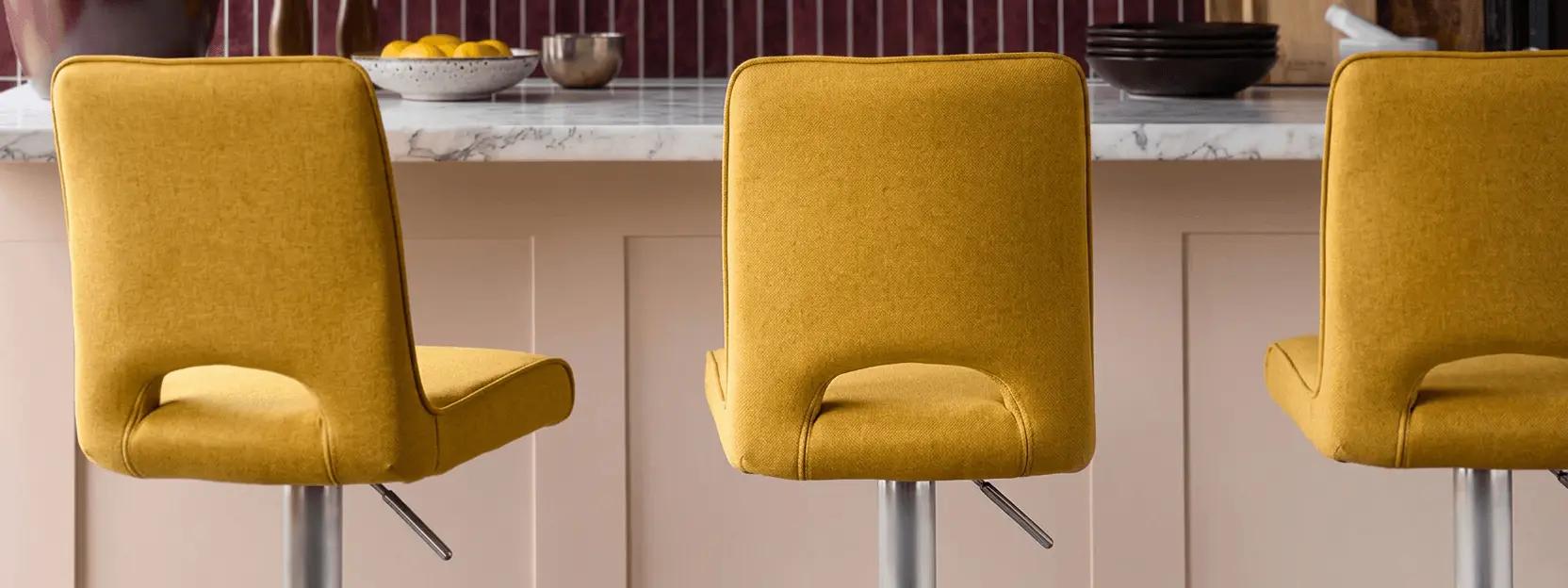 Three yellow bar stools with metal legs at a marble countertop, with bowls of fruit and dishes in the background.