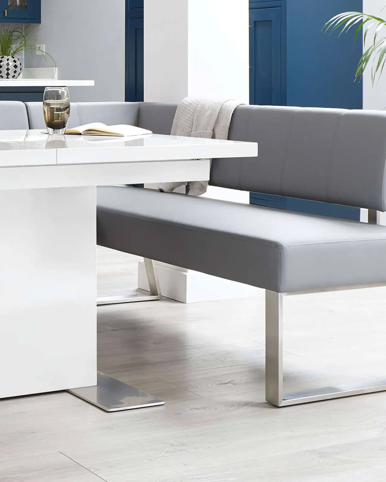 Modern dining area with white table and gray bench seating, featuring a glass and open book. Blue cabinets and a plant in the background.
