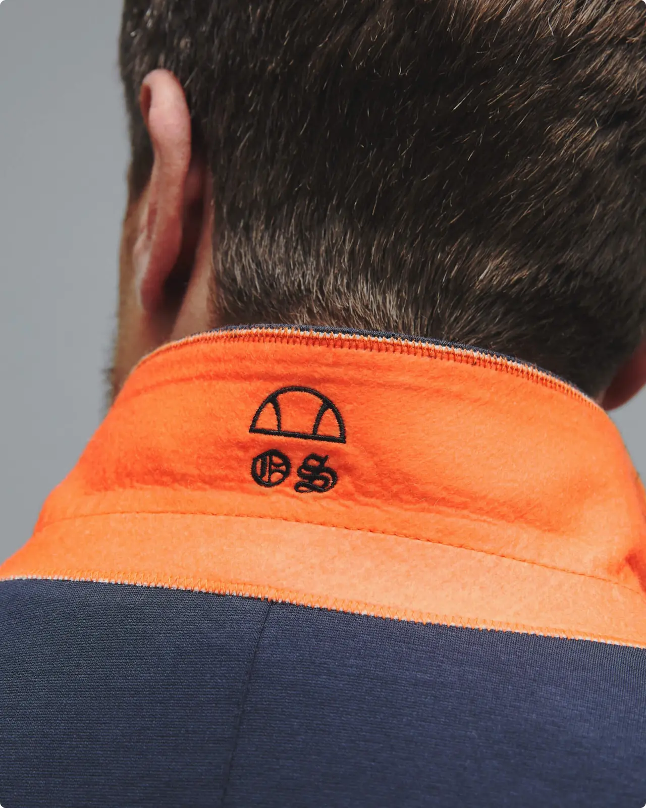 Close-up of a man’s back wearing an orange and navy jacket with embroidery featuring a semi-circle and “06” symbol on the collar.