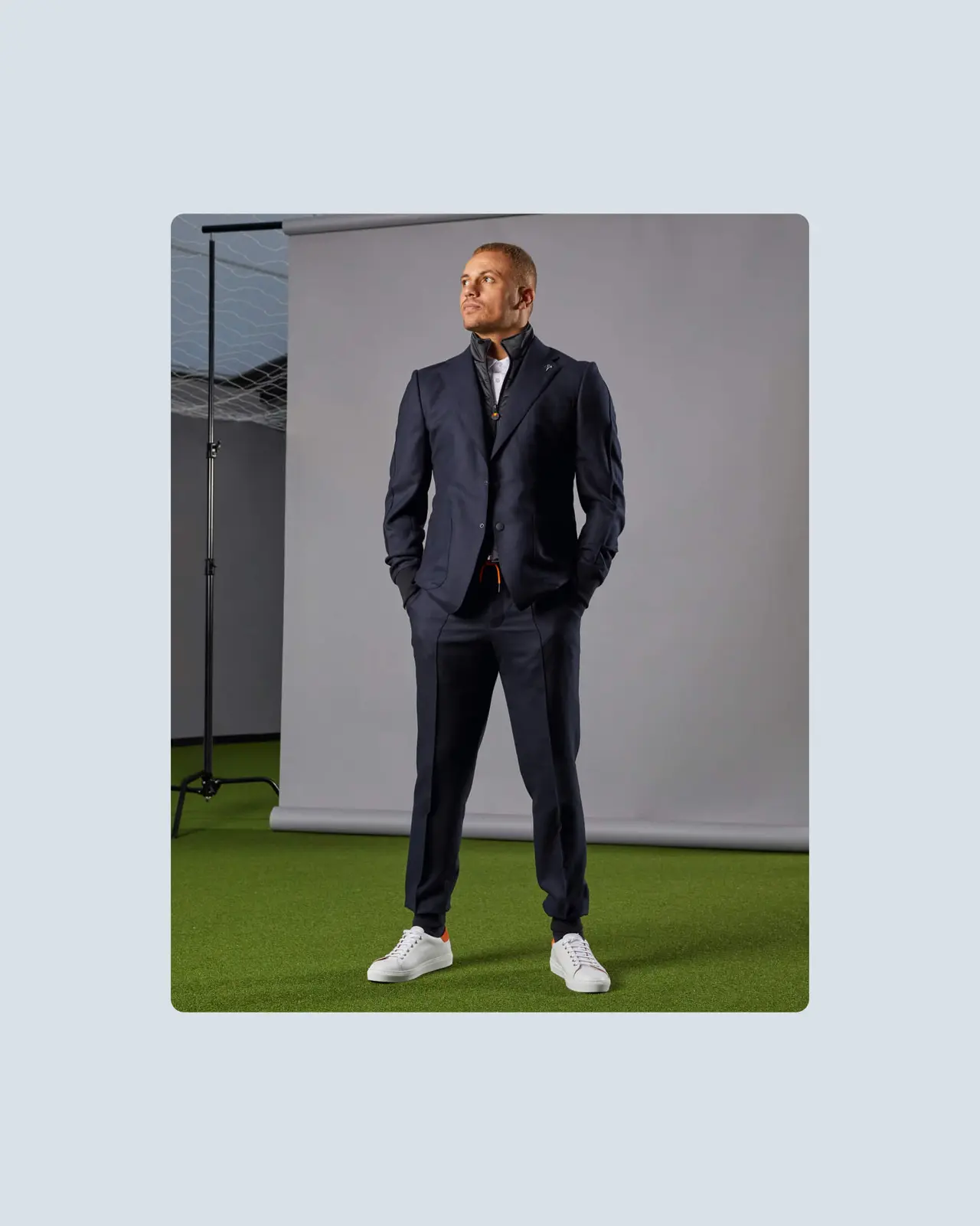 Wes Brown in a navy suit and white trainers stands confidently on green turf with a gray backdrop behind him.