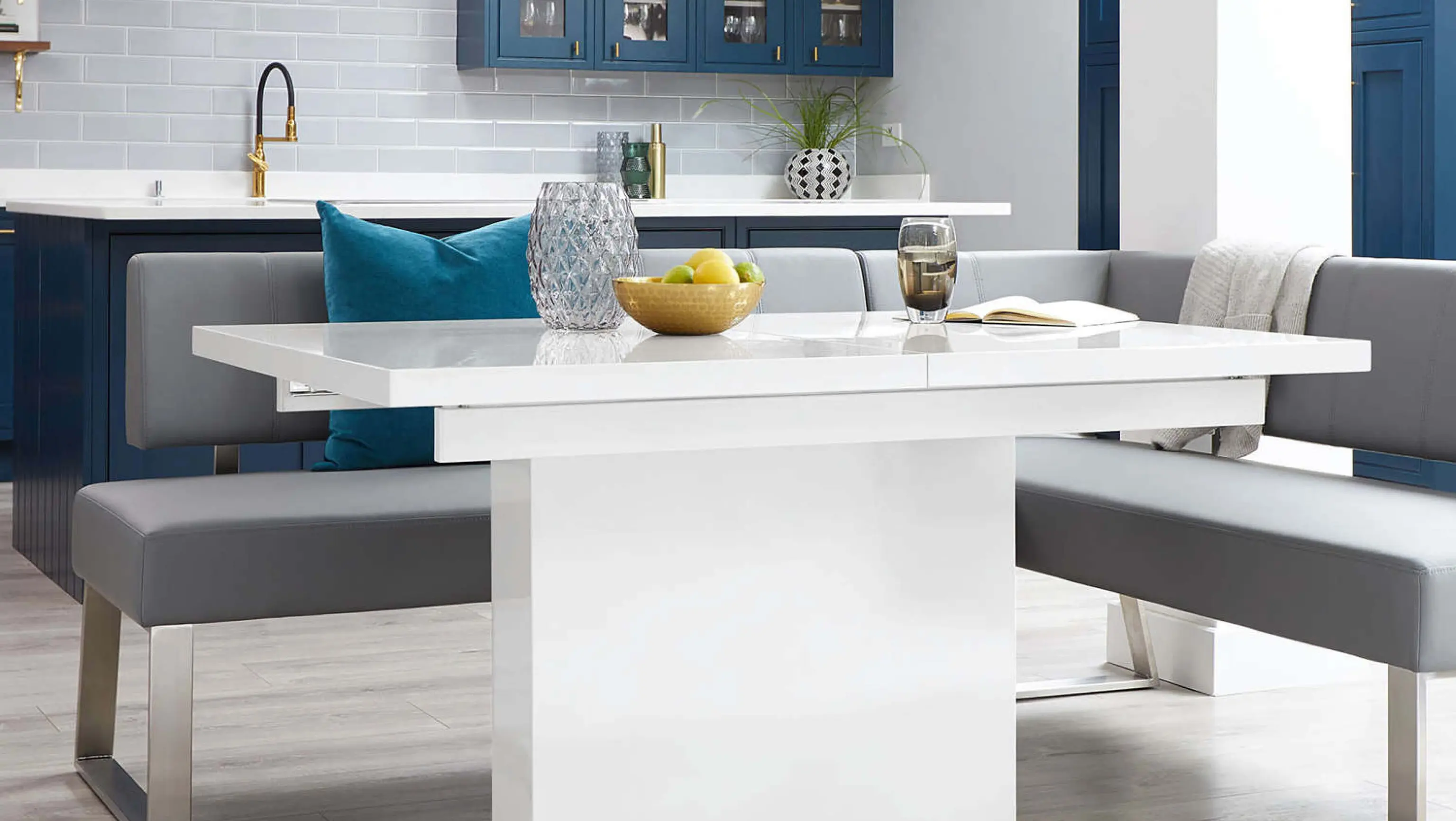 Modern dining area with a white table, gray benches, a bowl of green apples, a glass, and a book. Blue cabinets and a kitchen in the background.
