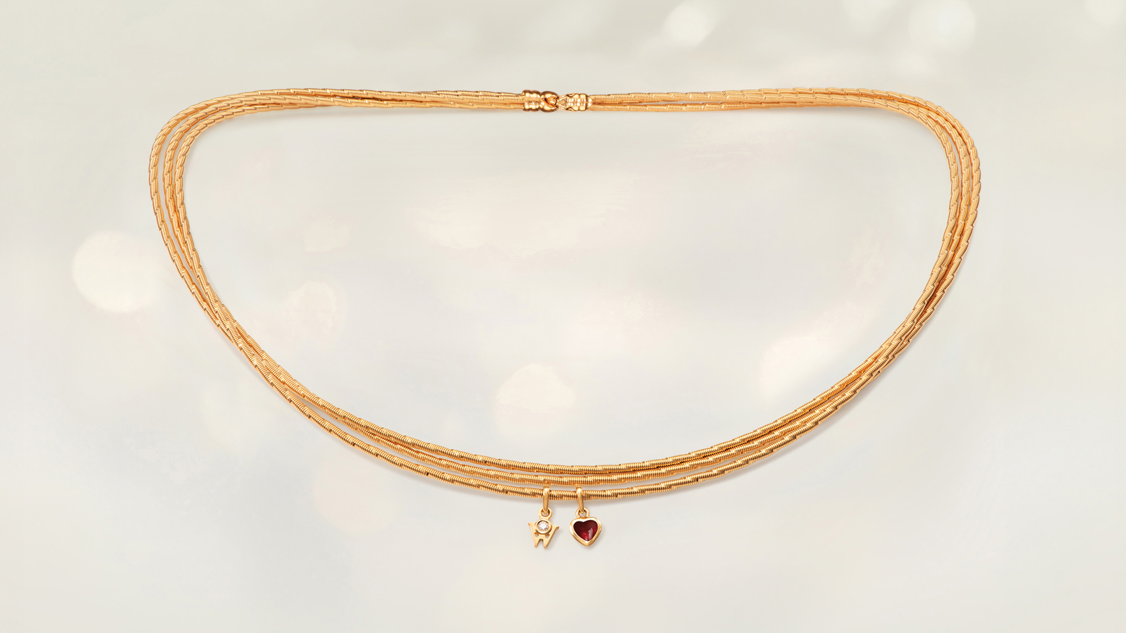 Das Collier MEIN HERZ Trio aus seidenweichem 18-karätigem Gold mit einem romantischen Herz gefüllt mit Wellendorff-Kaltemaille. Auf grauem Mood Hintergrund.