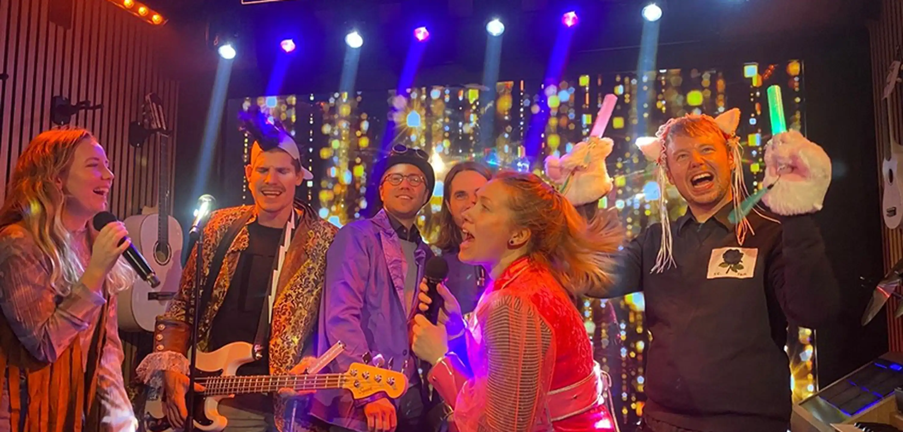 A lively group sings karaoke on stage with colorful lights and a sparkling backdrop, creating a festive atmosphere.