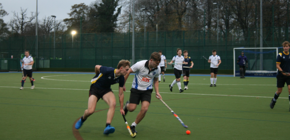 Two field hockey players battle for an orange ball on a green artificial turf pitch, teammates and a goalkeeper are behind them