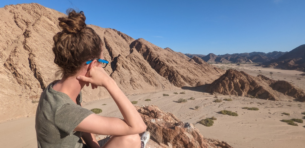 Ellie Harris sits on a rocky outcrop, looking out over a sunny, arid mountain range, in Namibia, Africa.