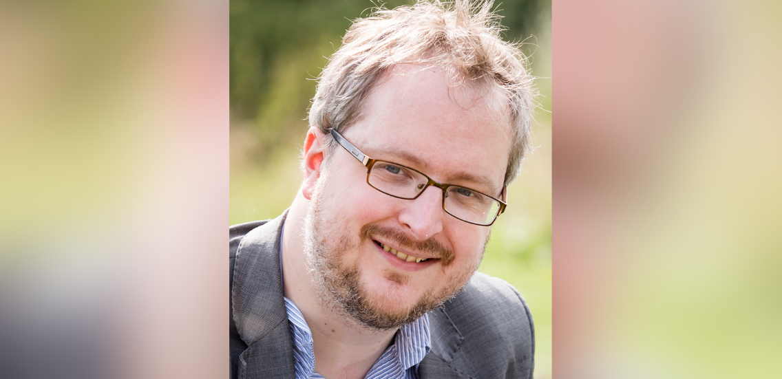 A professional outdoor headshot of a male academic. He is smiling, wearing glasses, a shirt, and a blazer.