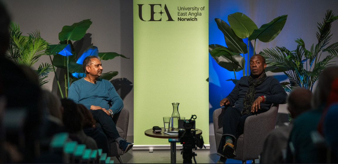 Two people seated in conversation on stage during a lecture at UEA with a crowd, in front of a green UEA banner.