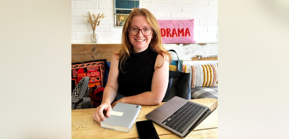 A student smiles while studying in a Norwich café, with her laptop and research materials on the table.