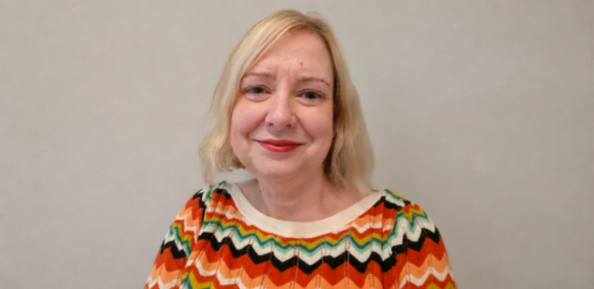 A professional headshot of a female academic with blonde hair, smiling while wearing a colourful chevron-patterned jumper against a plain wall.
