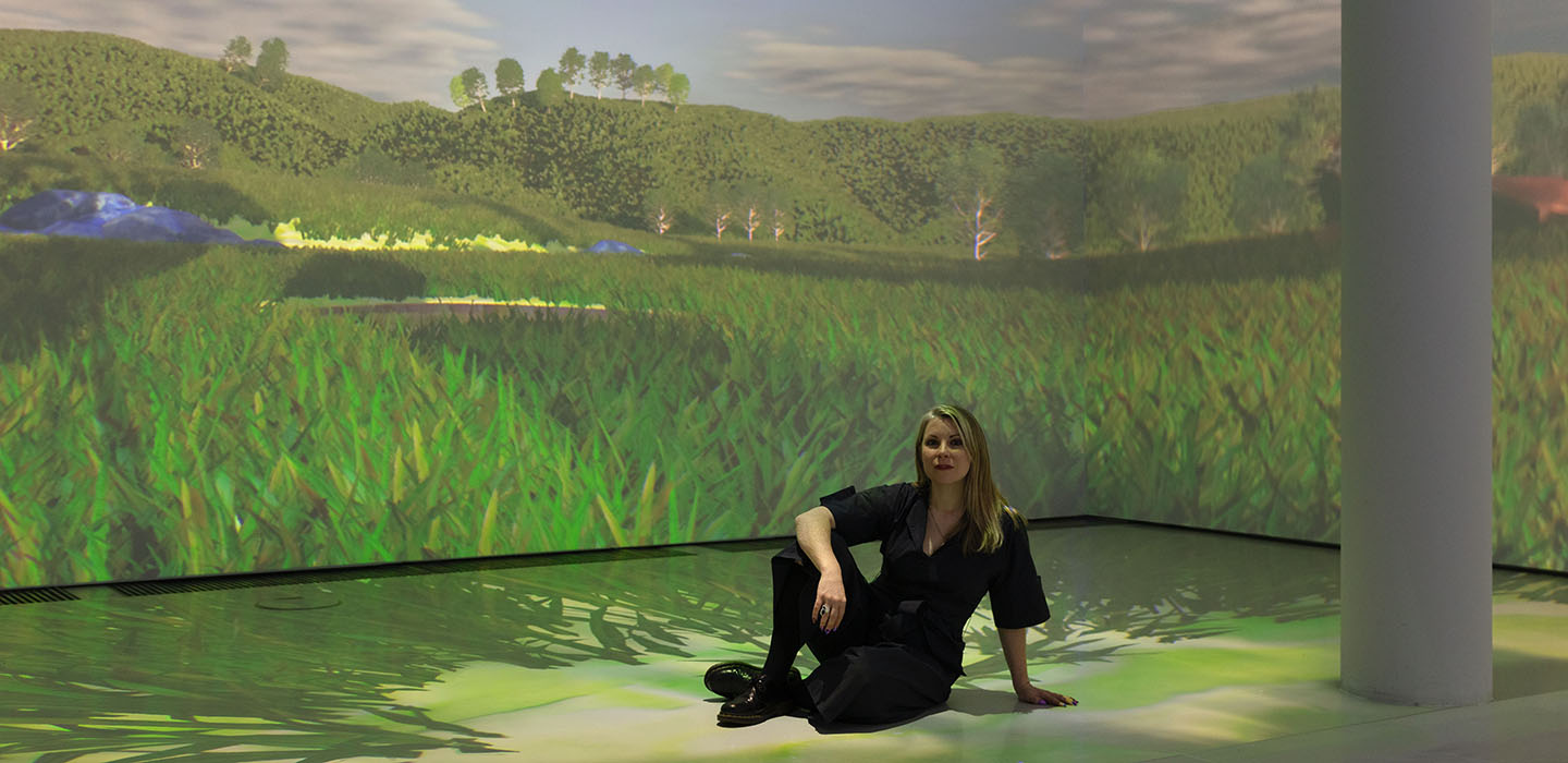 Libby Heaney sits cross-legged within an immersive projection of a virtual grassy landscape.