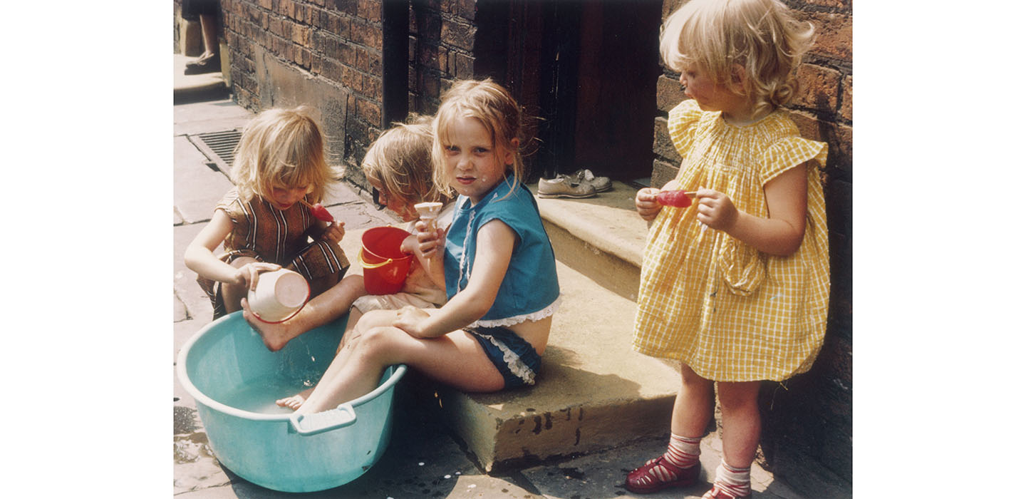 A colour photograph of four children playing on pavement, while they eat ice cream and lollies.