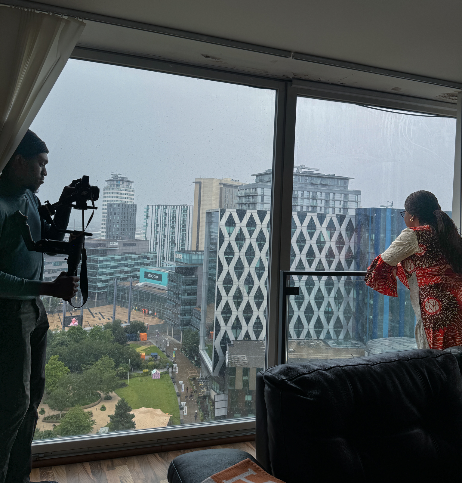 Betty Ajele is wearing colorful attire and looking out a large window at a cityscape on the right, while Ayoola films her with a camera on a stabiliser