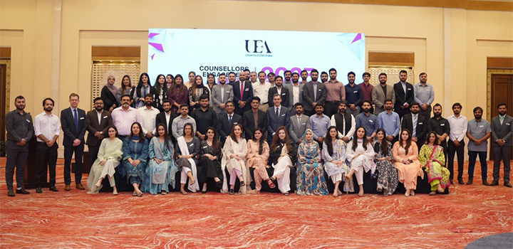 A large group of people, dressed formally, pose for a group photo in a spacious room with a UEA presentation screen behind them