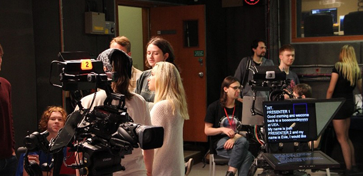 A busy TV studio with cameras, a teleprompter, and people interacting, preparing for a broadcast.
