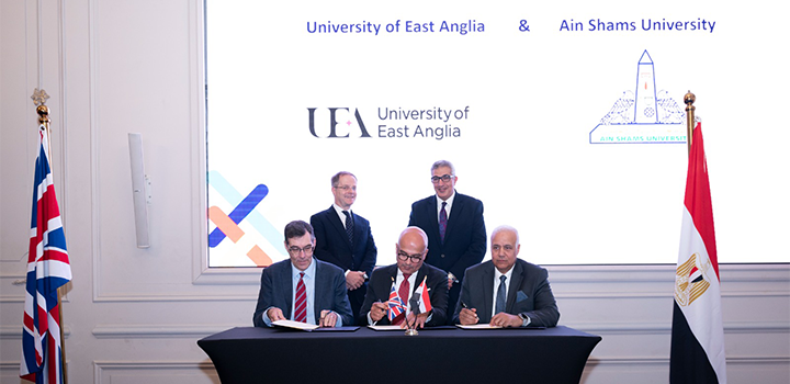 Three men are seated at a black table signing documents, while two other men stand behind them. Two flags are on either side of them, and a white screen behind them.
