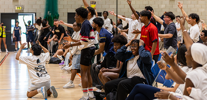 A group of enthusiastic spectators cheer and react energetically at an indoor sports event, with some seated and others standing
